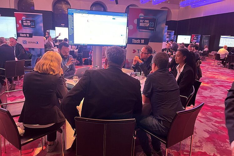 Teilnehmer sitzen an einem runden Tisch während einer ITIL-Tool-Meetup-Session, diskutieren und betrachten eine Präsentation auf einem großen Bildschirm.
