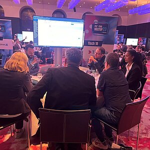 Teilnehmer sitzen an einem runden Tisch während einer ITIL-Tool-Meetup-Session, diskutieren und betrachten eine Präsentation auf einem großen Bildschirm.