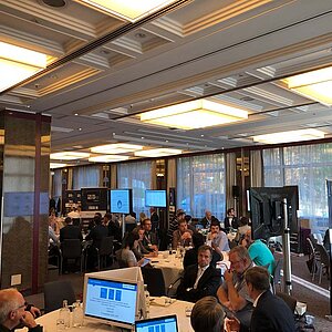 Teilnehmer sitzen an einem runden Tisch während einer Tool-Meetup-Session, diskutieren und betrachten eine Präsentation auf einem großen Bildschirm.