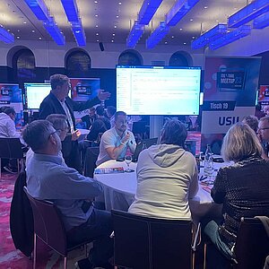 Teilnehmer sitzen an einem runden Tisch während einer Tool-Meetup-Session, diskutieren und betrachten eine Präsentation auf einem großen Bildschirm.