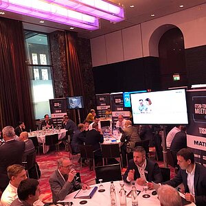 Teilnehmer des ITSM-Tool MeetUp sitzen an runden Tischen in einem modernen Konferenzsaal mit großen Bildschirmen und LED-Deckenbeleuchtung.
