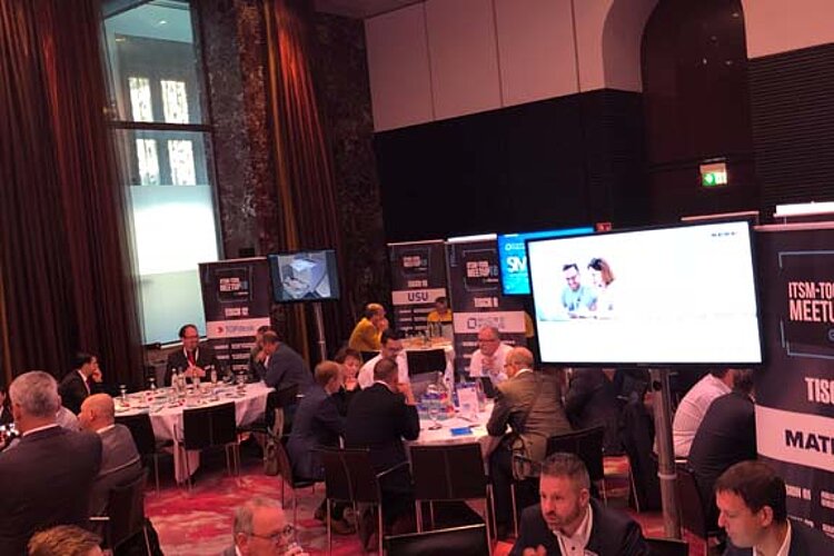 Teilnehmer des ITSM-Tool MeetUp sitzen an runden Tischen in einem modernen Konferenzsaal mit großen Bildschirmen und LED-Deckenbeleuchtung.