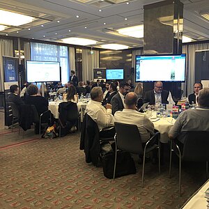 Teilnehmer des ITSM-Tool MeetUp18 sitzen an runden Tischen und diskutieren mit Herstellern, während Präsentationen auf großen Bildschirmen gezeigt werden.