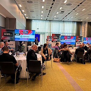 Teilnehmer des SERVIEW TOOL MEETUP24 sitzen an runden Tischen und diskutieren mit Herstellern, während Präsentationen auf großen Bildschirmen gezeigt werden.