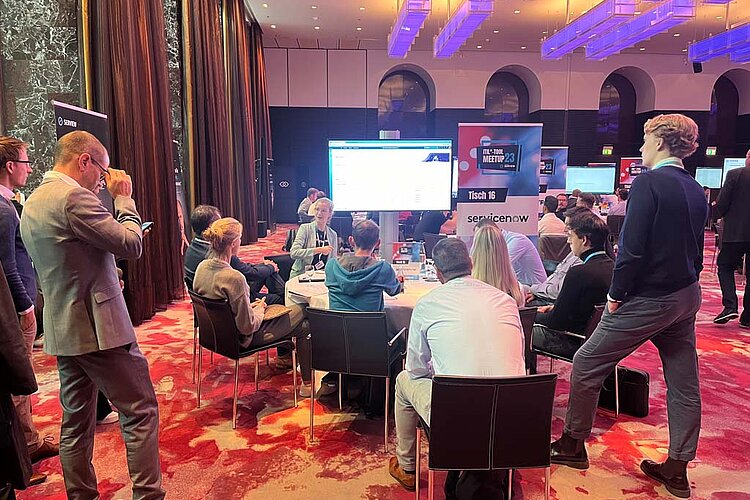 Teilnehmer sitzen an einem runden Tisch während einer Tool-Meetup-Session, diskutieren und betrachten eine Präsentation auf einem großen Bildschirm.
