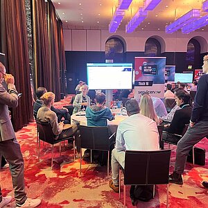 Teilnehmer sitzen an einem runden Tisch während einer Tool-Meetup-Session, diskutieren und betrachten eine Präsentation auf einem großen Bildschirm.