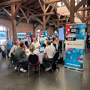 Teilnehmer sitzen an einem runden Tisch während einer Tool-Meetup-Session, diskutieren und betrachten eine Präsentation auf einem großen Bildschirm.