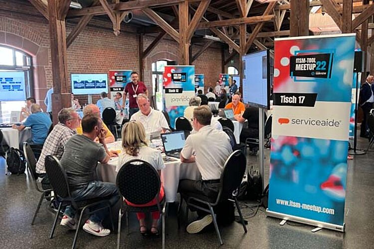 Teilnehmer sitzen an einem runden Tisch während einer Tool-Meetup-Session, diskutieren und betrachten eine Präsentation auf einem großen Bildschirm.