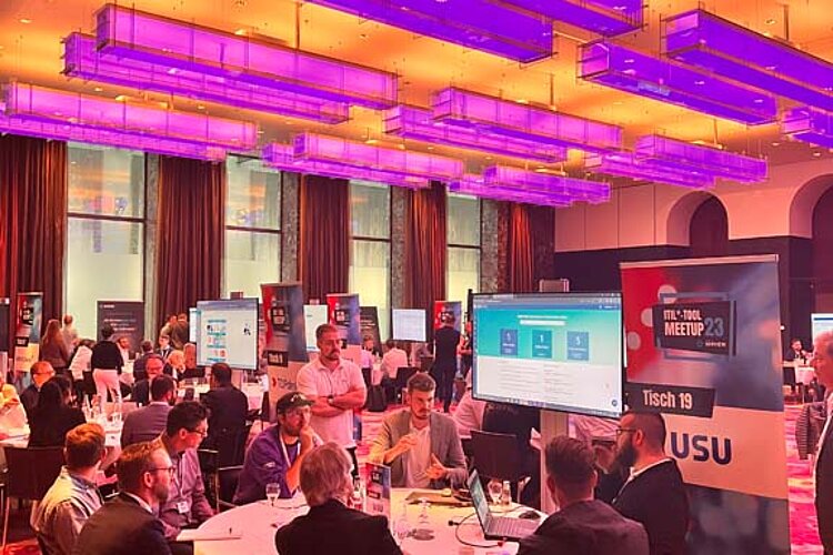 Teilnehmer sitzen an einem runden Tisch während einer Tool-Meetup-Session, diskutieren und betrachten eine Präsentation auf einem großen Bildschirm.