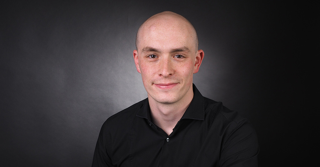 Porträt eines Mannes mit schwarzem Hemd vor einem dunklen Hintergrund, der freundlich in die Kamera lächelt.